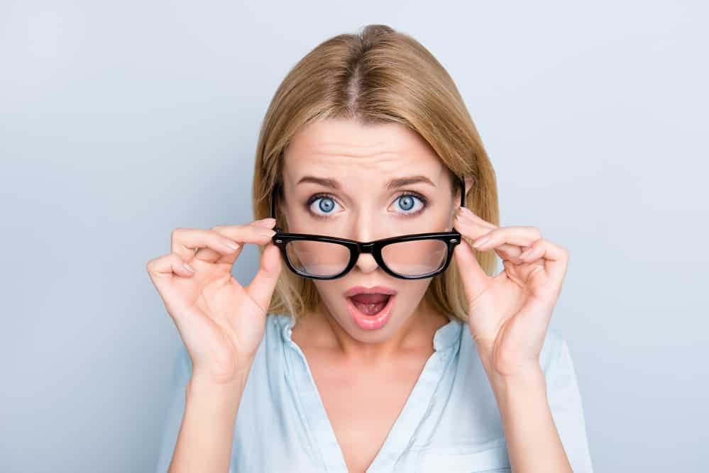 Young woman wearing glasses, holding glasses below eyes with shocked expression