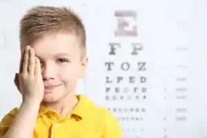 Boy holding a hand over one eye, in front of eye exam poster