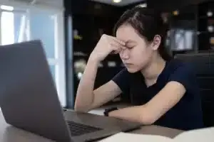 Young woman in front of desktop with eyes closed, frustrated with dry eyes