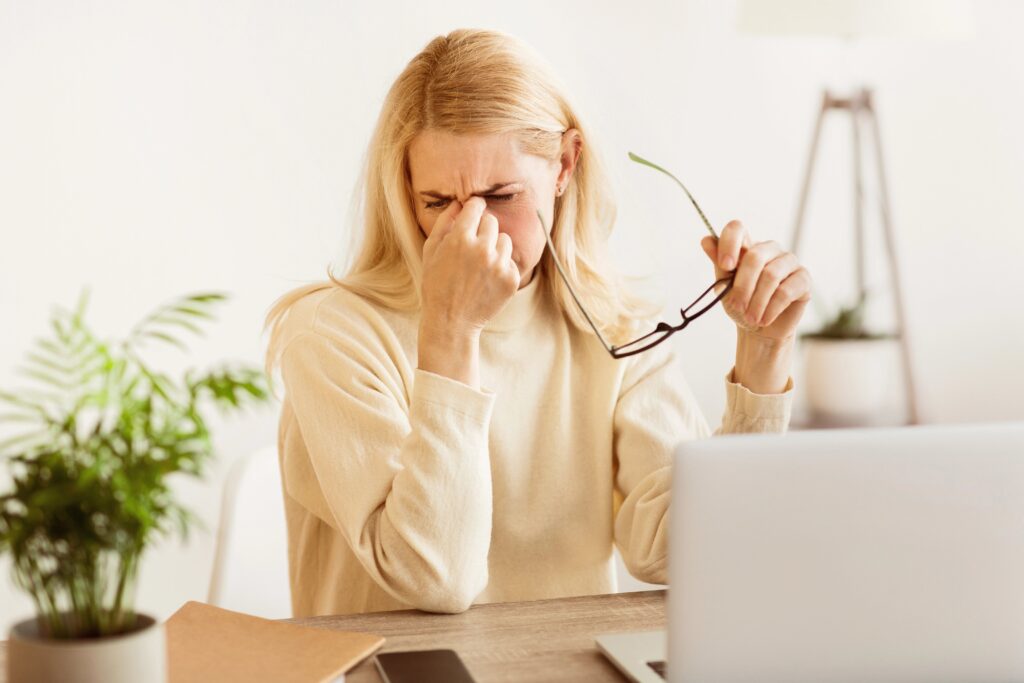 Woman in front of computer, holding glasses, rubbing eyes due to eye strain